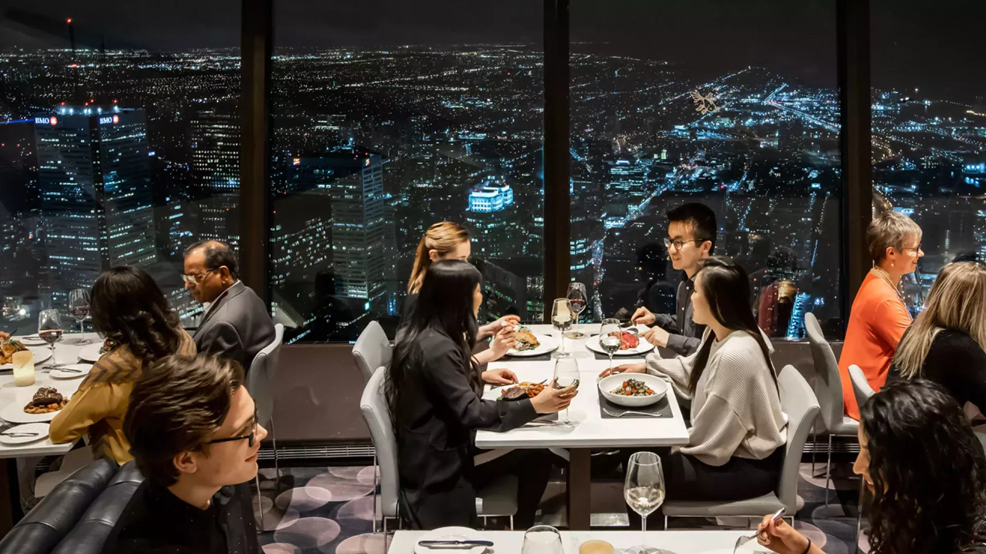 Plusieurs personnes assises à des tables à manger avec les lumières de la ville en arrière-plan.