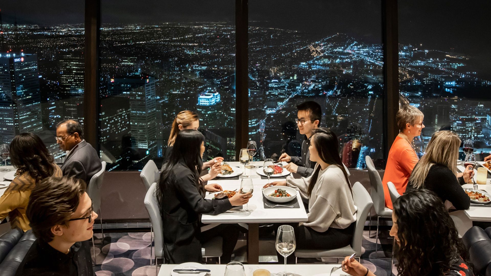 Plusieurs personnes assises à des tables à manger avec les lumières de la ville en arrière-plan.