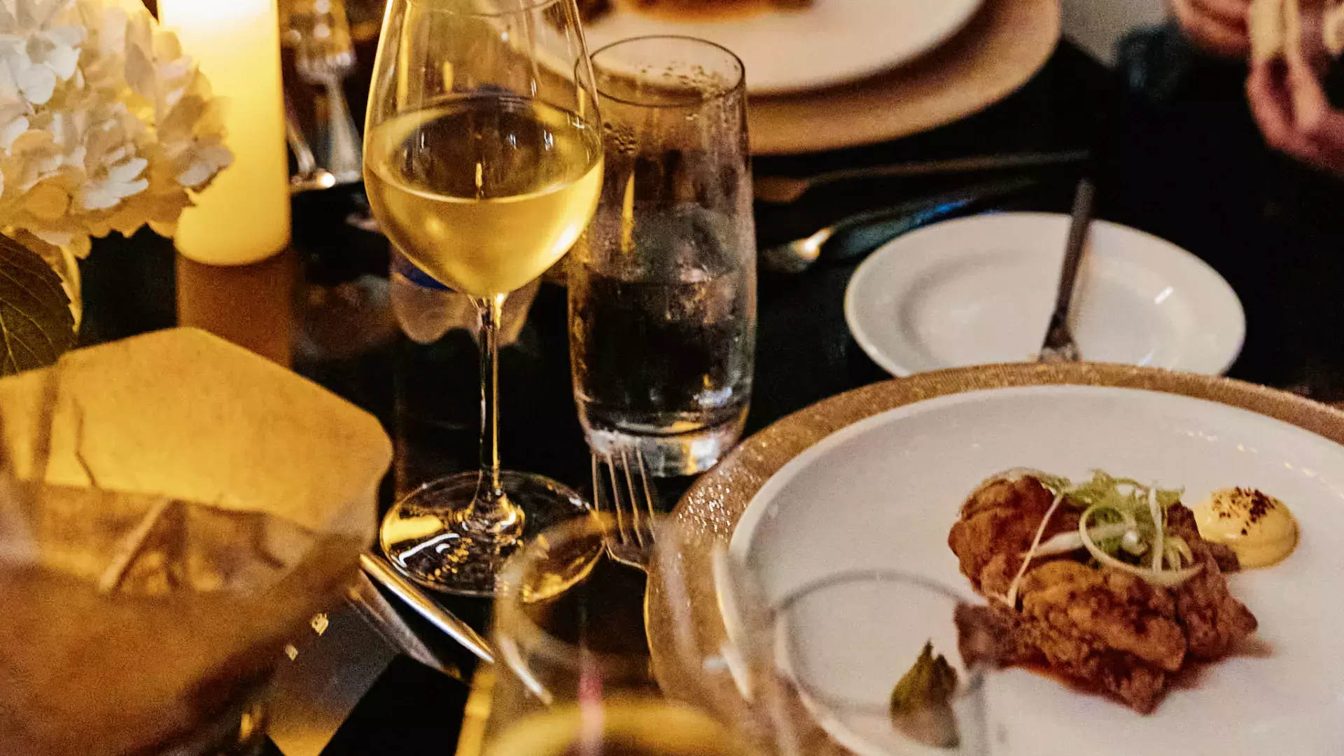Gros plan sur de multiples objets (assiette de nourriture, verre de vin, petit plat d'accompagnement, couverts, fleurs) dispersés sur une table le soir, éclairée par une bougie.