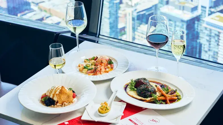 Le menu dégustation Expérience canadienne au restaurant 360 de la Tour CN, composé de plusieurs plats inspirés des saveurs canadiennes, dont un plat de fruits de mer, une viande braisée accompagnée de légumes de saison, un dessert et une bouchée gastronomique, le tout accompagné de vins rouge, blanc et mousseux. La table est installée près de baies vitrées offrant une vue panoramique sur le centre-ville de Toronto depuis la Tour CN.
