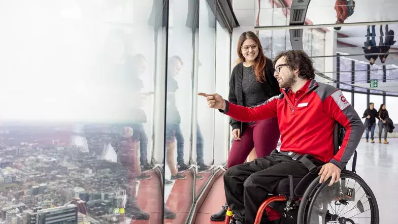 Un membre de l'équipe de la Tour CN, un homme de race blanche, portant une chemise rouge et se déplaçant en fauteuil roulant, montre quelque chose par la fenêtre pleine hauteur à une cliente, une femme de race blanche, qui se tient derrière lui au Niveau d'observation principal.