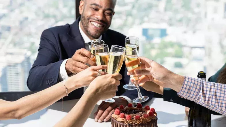 Homme de race noire trinquant avec des amis au 360, avec la vue de Toronto à l'arrière et un gâteau au chocolat sur la table.
