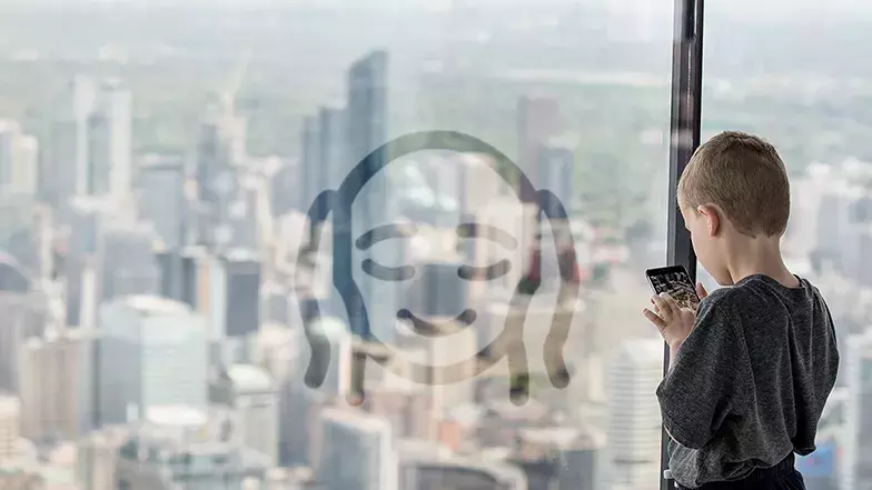 Un jeune garçon blanc se tient au Niveau d'observation principal de la Tour CN, contre les murs de fenêtres allant du sol au plafond, face à la ville et tient un téléphone intelligent. Nous voyons l'icône des Expériences sensorielles réduites en surimpression sur l'image