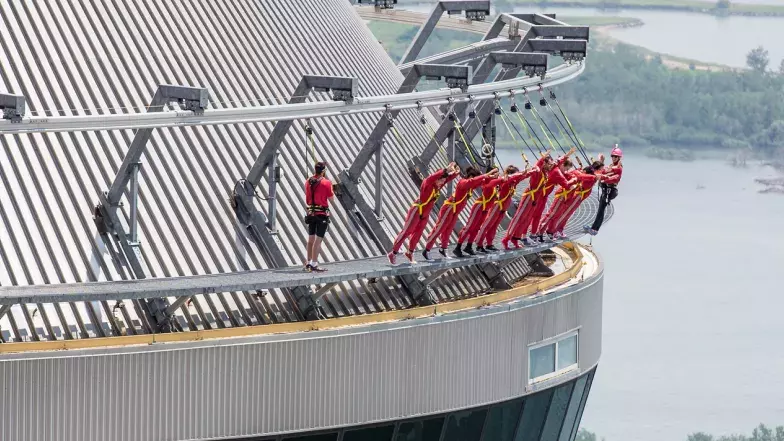 Un groupe de personnes à l'extérieur de la tour (L'HAUT-DA CIEUX). Ils portent des harnais reliés à des lignes de sécurité et se penchent vers l'avant par-dessus le bord de la tour, les bras tendus.
