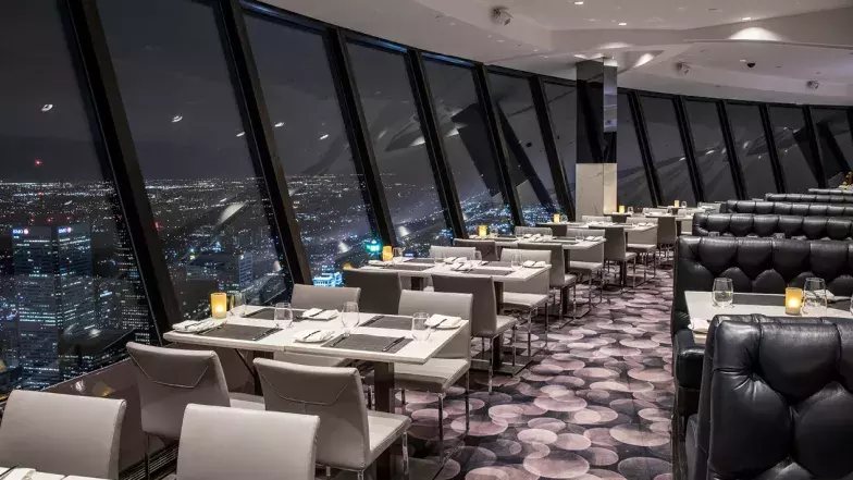 La fenêtre du restaurant montre Toronto la nuit. Le long de la fenêtre, il y a des tables et des chaises. De l'autre côté de l'allée, il y a des cabines. Sur les tables, des bougies sont allumées. Il n'y a personne dans cet espace.