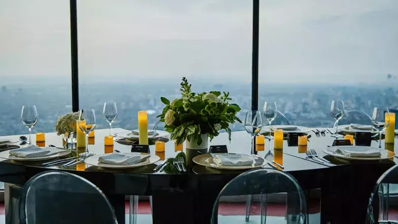 Vue d'une table en serpentin installée au niveau d'observation principal de la Tour CN. Au centre, un gros bouquet de fleurs blanches, entouré d'assiettes vides, de bougies et d'autres fleurs.