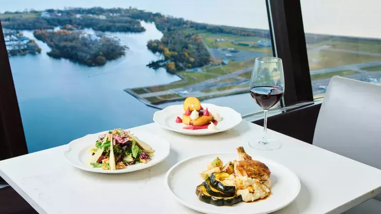 Vue de trois assiettes posées sur une table en marbre blanc au restaurant 360. En arrière-plan, un verre de vin rouge et les fenêtres du restaurant donnant sur le lac Ontario.