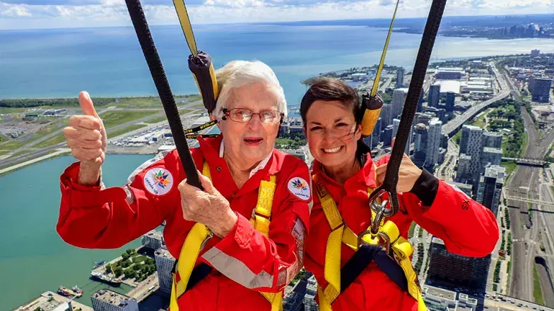 Deux personnes souriant et levant le pouce, avec la ville de Toronto en contrebas, par une journée d'été claire mais nuageuse.