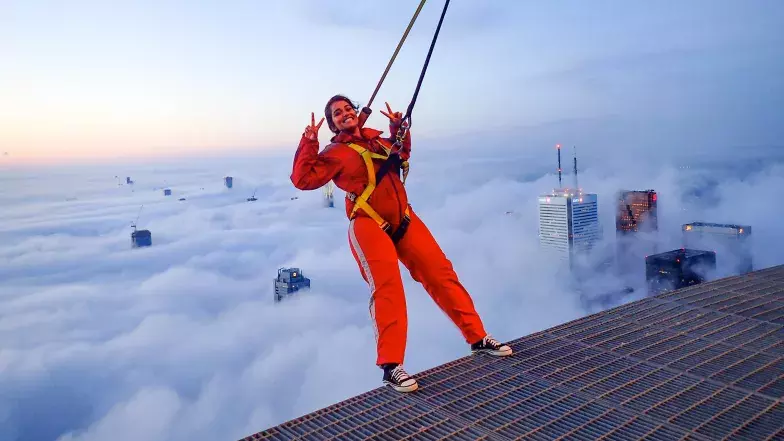 Une personne dans un harnais se penche en arrière au-dessus de la promennade. Les nuages sont en dessous.