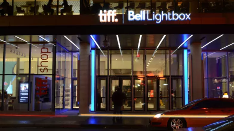 Mur de verre avec des portes en verre. Deux voitures passent et une personne se tient devant le bâtiment. L'enseigne lumineuse au-dessus des portes indique Tiff Bell Lightbox