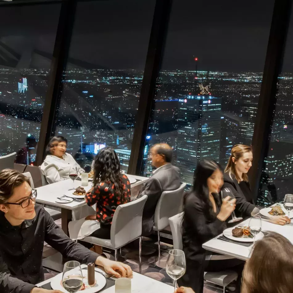 6 personnes assises à 3 tables en train de dîner. Lumières de la ville de Toronto la nuit en arrière-plan
