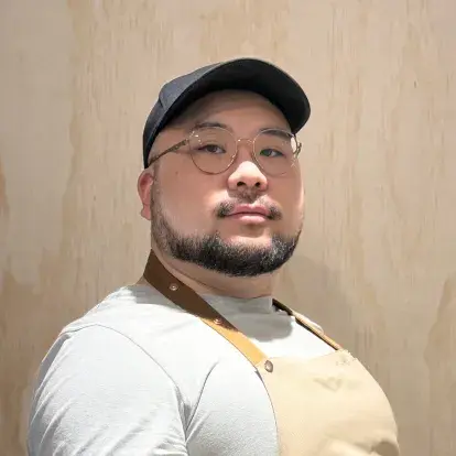 Portrait de Jin Ke Wang faisant face à la caméra à l’intérieur, portant des lunettes, une casquette et un tablier, sur un fond en panneaux de bois clair.