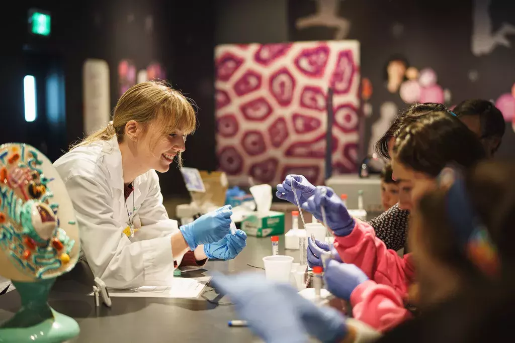 Une femme en blouse de laboratoire assistant les enfants dans le cadre d’une expérience scientifique au Centre des sciences de Montréal.