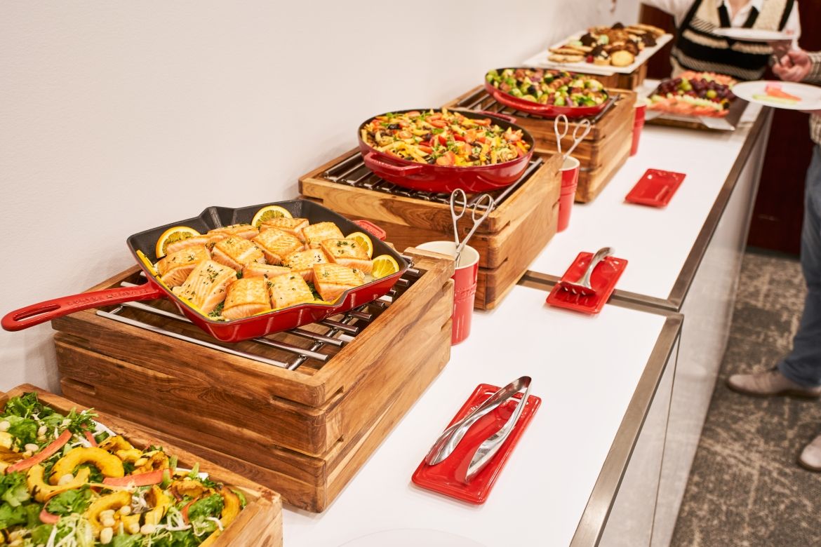 Detailed view of one end of the food table. Dishes are presented in red cast iron skillets, each atop a wood block with a metal grill. Tongs are available in a red cup next to each dish and serving utensils is placed on a red spoon rest in front of each dish. Dishes in this view are salmon, pasta and salad. 