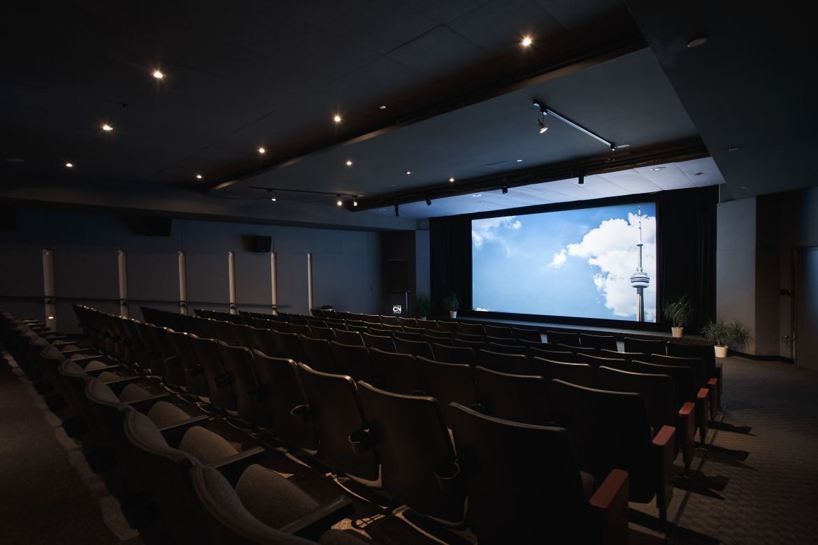 Large, darkened theatre room with seven rows of sixteen upholstered chairs facing a large screen.