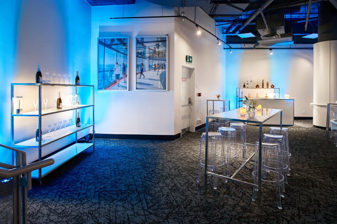 Large reception area lit in blue with a bar area and shelf displaying champagne glasses and bottles of champagne. There are several high-top tables with stools for seating. 