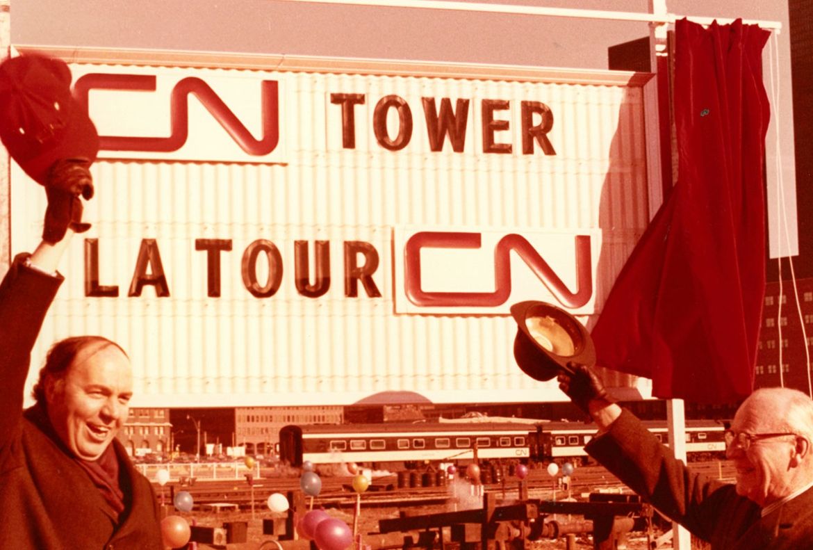 Deux hommes saluent avec leur chapeau en direction d’une affiche portant l’inscription « CN Tower » pour marquer le début de la construction de la Tour en février 1973.