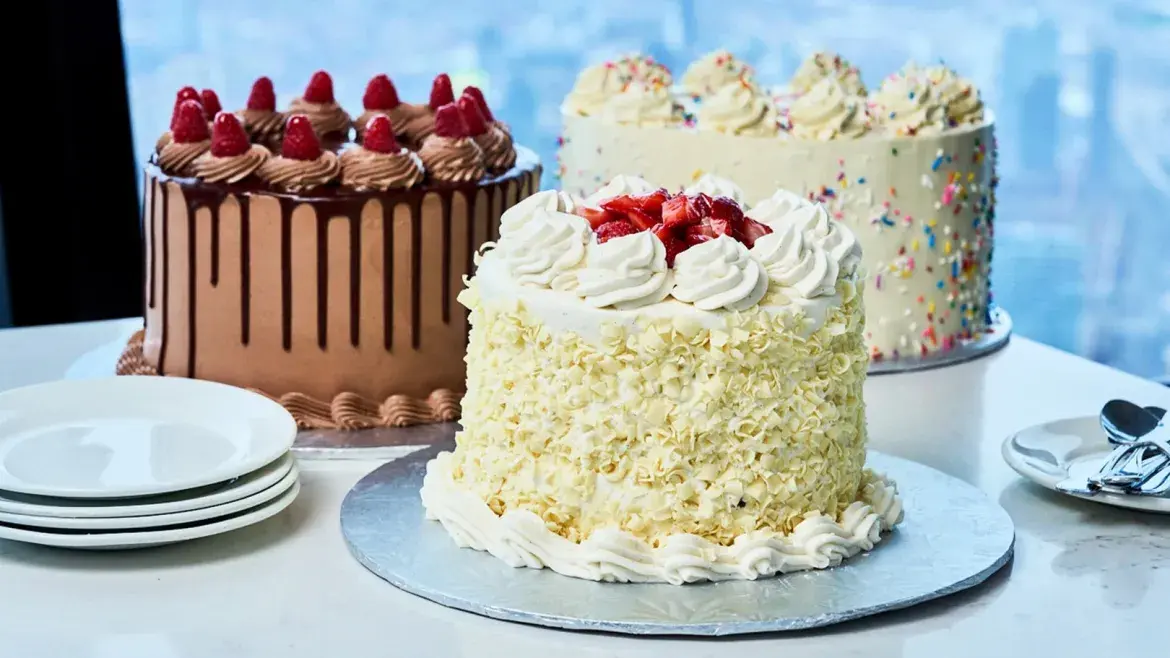 Trois gâteaux décorés présentés sur une table blanche au restaurant 360, comprenant des gâteaux au chocolat et à la vanille garnis de framboises, de fraises, de glaçage et de vermicelles, accompagnés d’assiettes à dessert et de cuillères, devant de grandes fenêtres inclinées avec vue sur la ville.
