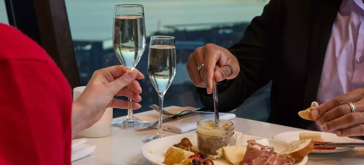 Un couple trinque joyeusement au champagne autour d'une table joliment dressée et ornée d'une planche de charcuterie. En arrière-plan, on peut voir la vue de Toronto au coucher du soleil à travers les fenêtres.
