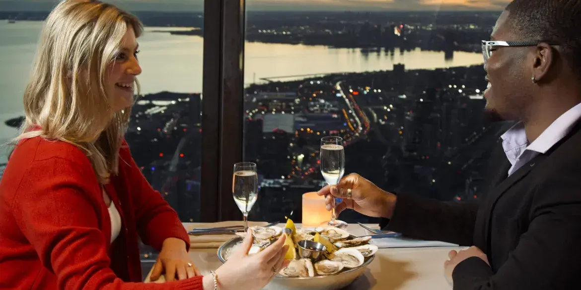 Un couple déguste des huîtres fraîches et du champagne au restaurant 360. En arrière-plan, on peut voir la vue de Toronto la nuit à travers les fenêtres.