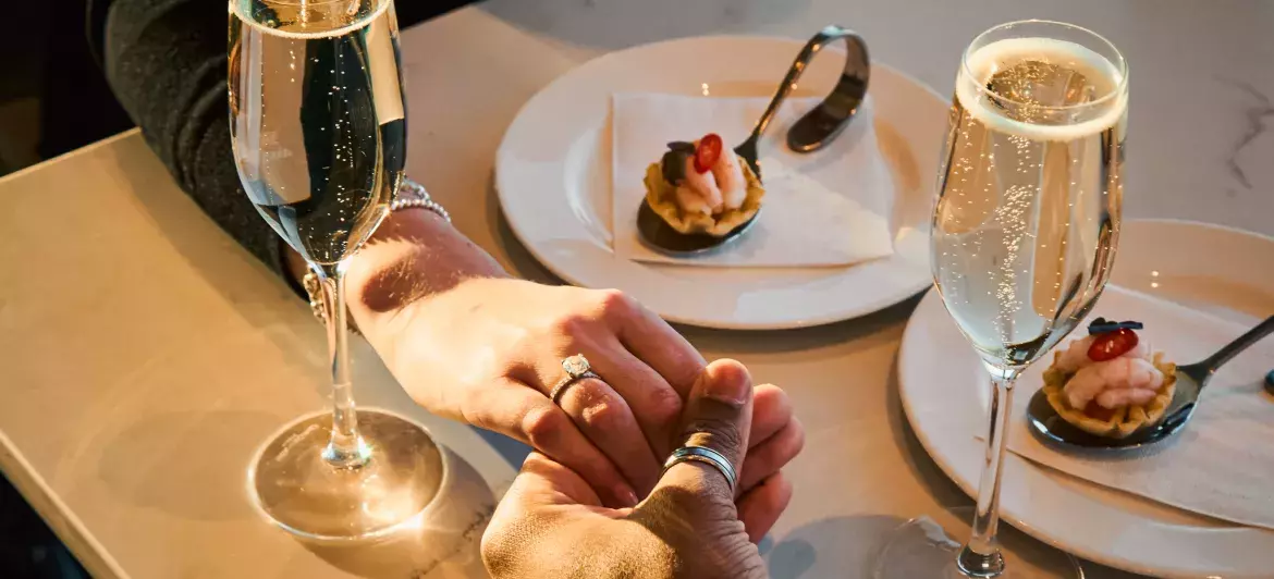 Deux mains entrelacées au-dessus de coupes de champagne, représentant un moment romantique partagé par un couple.