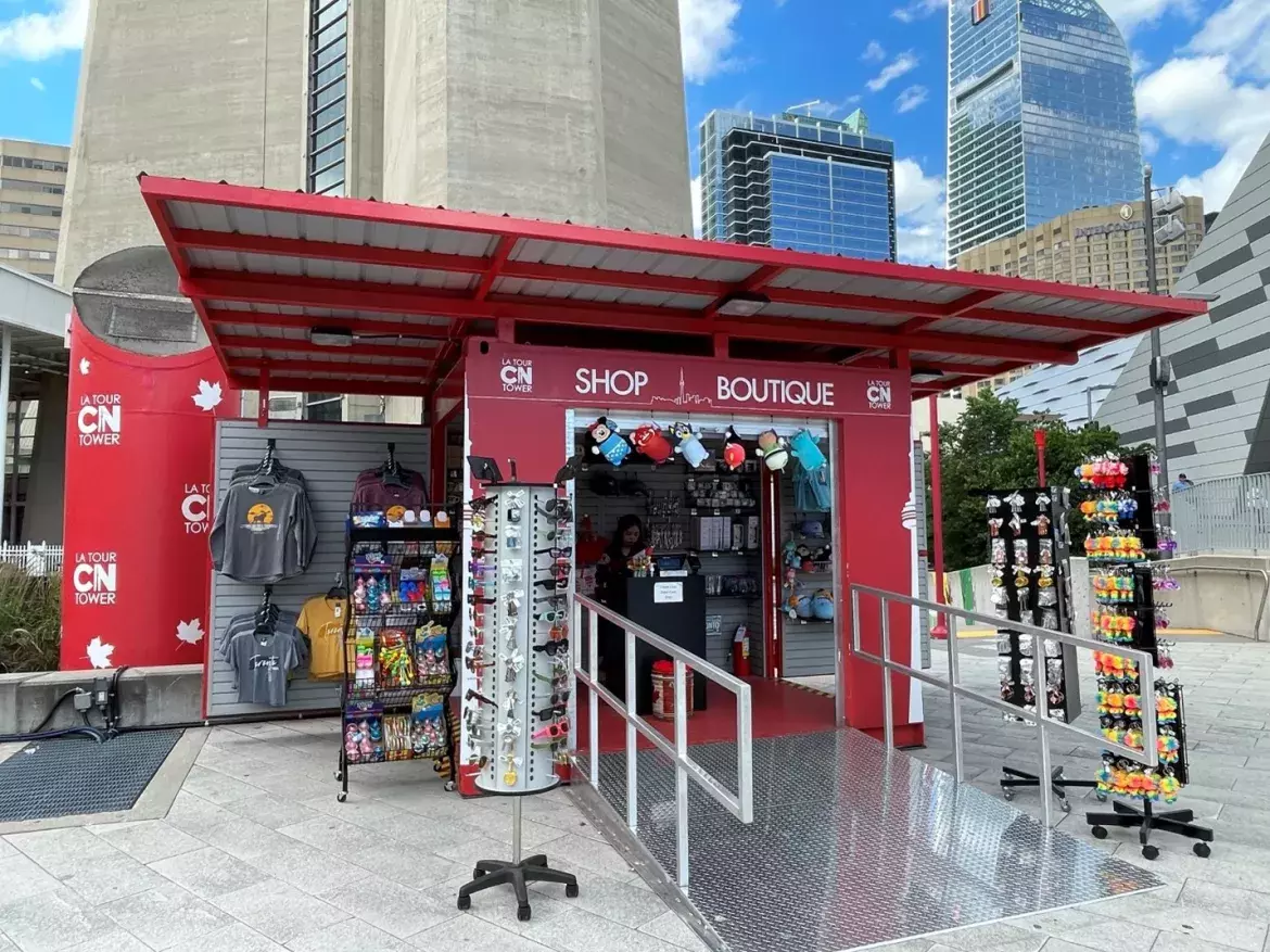 Kiosque extérieur rouge situé sur la Place de la Société immobilière du Canada, avec le logo de la Tour CN et les mots Shop/Boutique sur le portique. Des articles divers tels que des t-shirts, des porte-clés et des lunettes de soleil sont exposés sur des présentoirs à l'intérieur et autour du kiosque. 